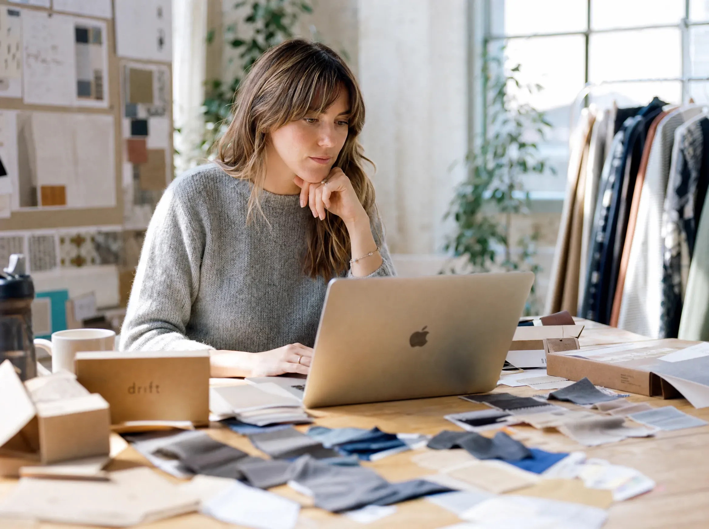 A woman in a grey knit sweater studies her laptop screen at a wooden desk scattered with folded fabric swatches and a branded shipping box.