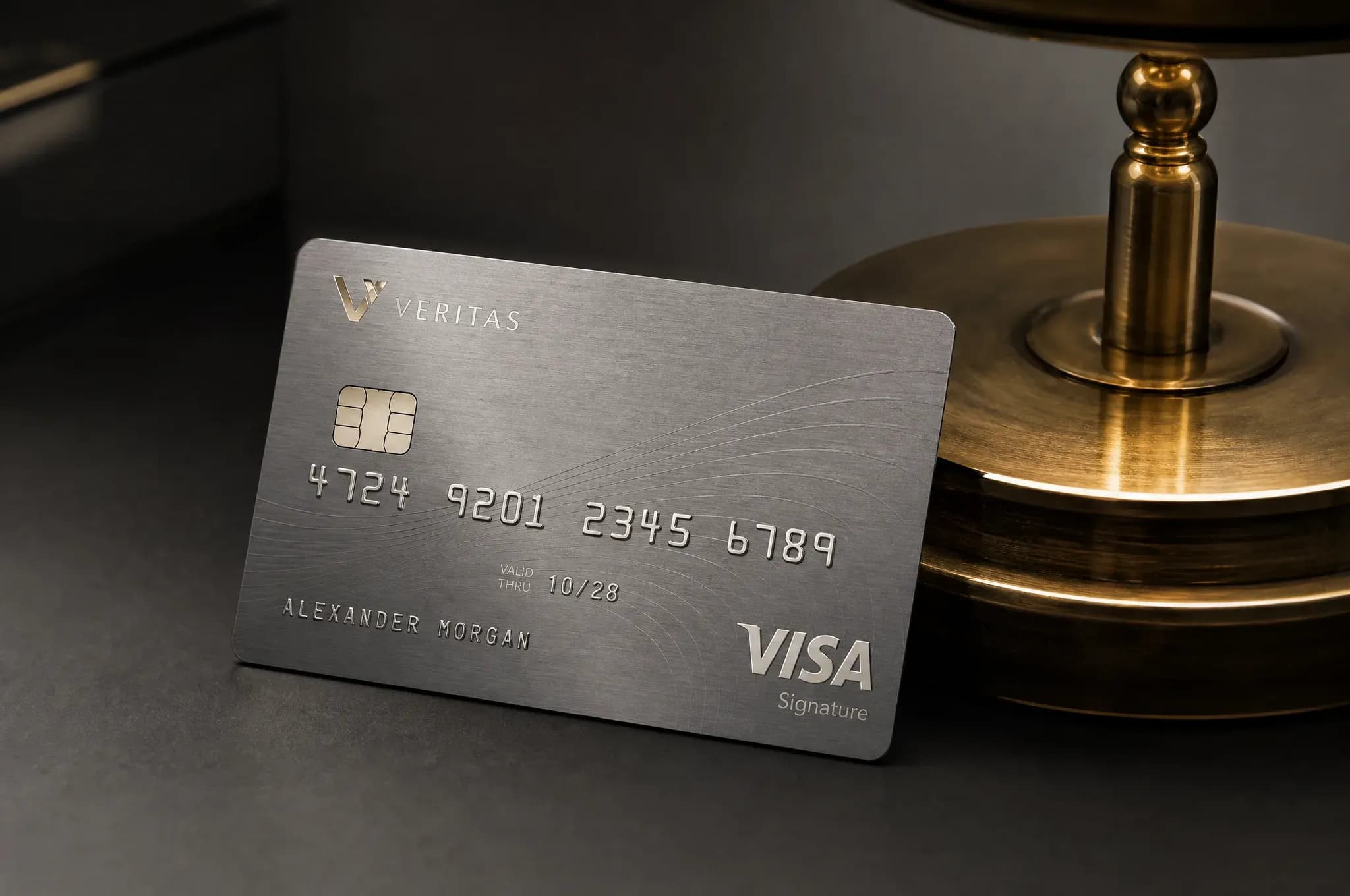 A brushed metal signature payment card, embossed with a cardholder name and chip, leans against a polished brass lamp base on a dark surface.