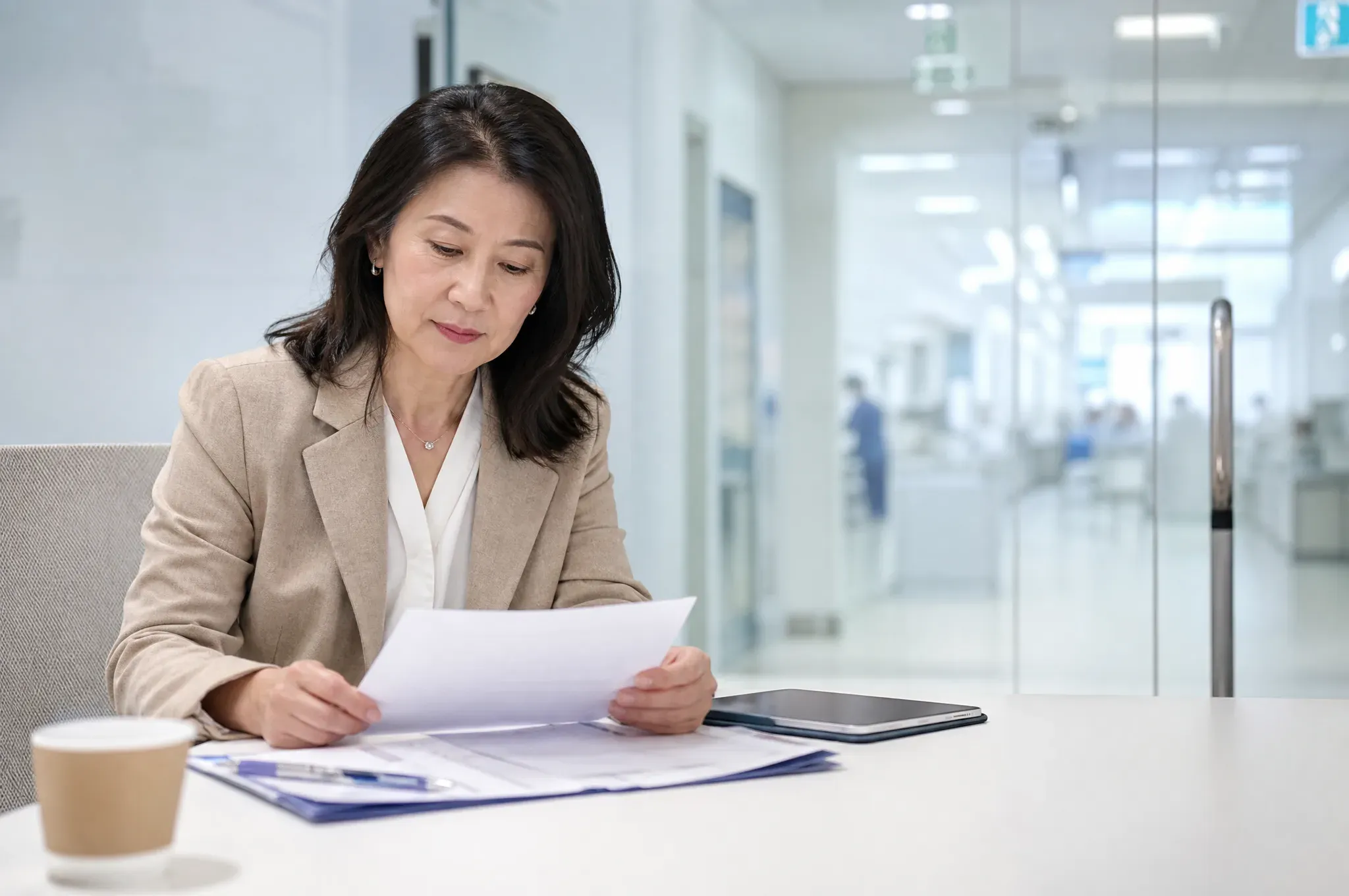 A woman in a beige blazer sits at a table reading a stack of papers, with a tablet beside her and a hospital corridor with a staff member in scrubs visible through the glass behind her.