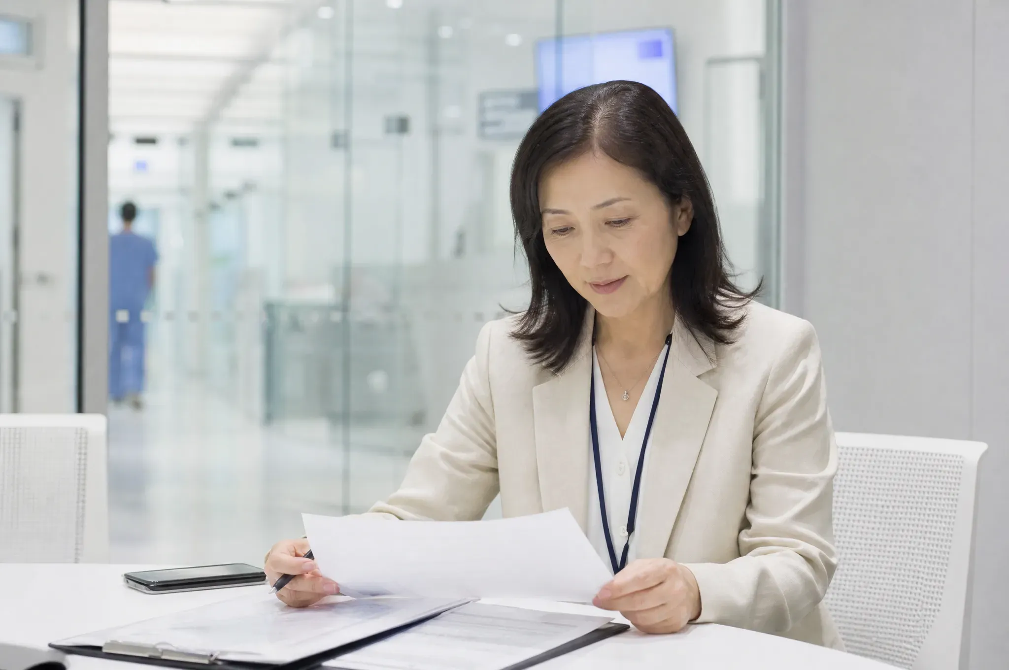 A woman in a cream blazer wearing a lanyard badge sits at a table, pen in hand, reviewing paperwork on a clipboard, with a hospital corridor and a staff member in scrubs visible behind her.