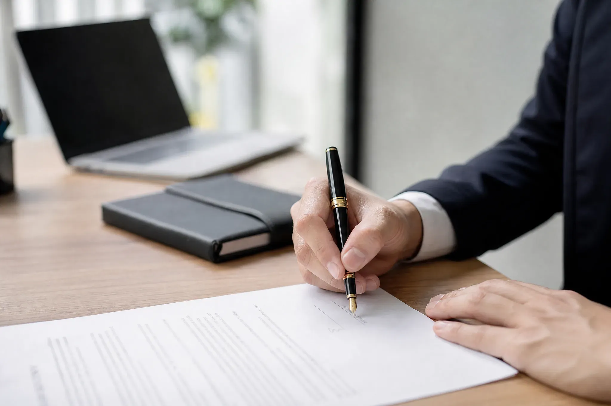A businessman in a navy suit signs a printed contract with a fountain pen at a wooden desk, with an open laptop and a black notebook nearby.