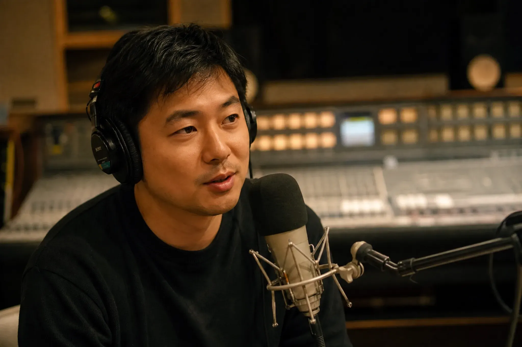 A man wearing headphones speaks into a studio microphone in a dimly lit recording studio with a large mixing console behind him.