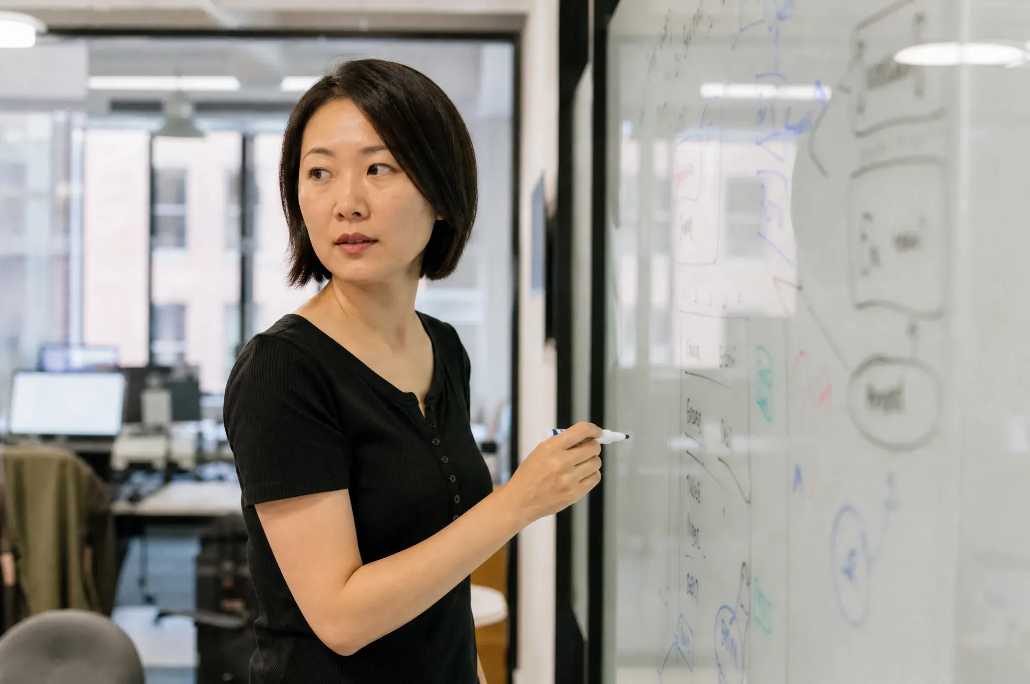 A SaaS designer with a bob haircut writes on a glass whiteboard covered in app wireframe sketches and flowcharts.