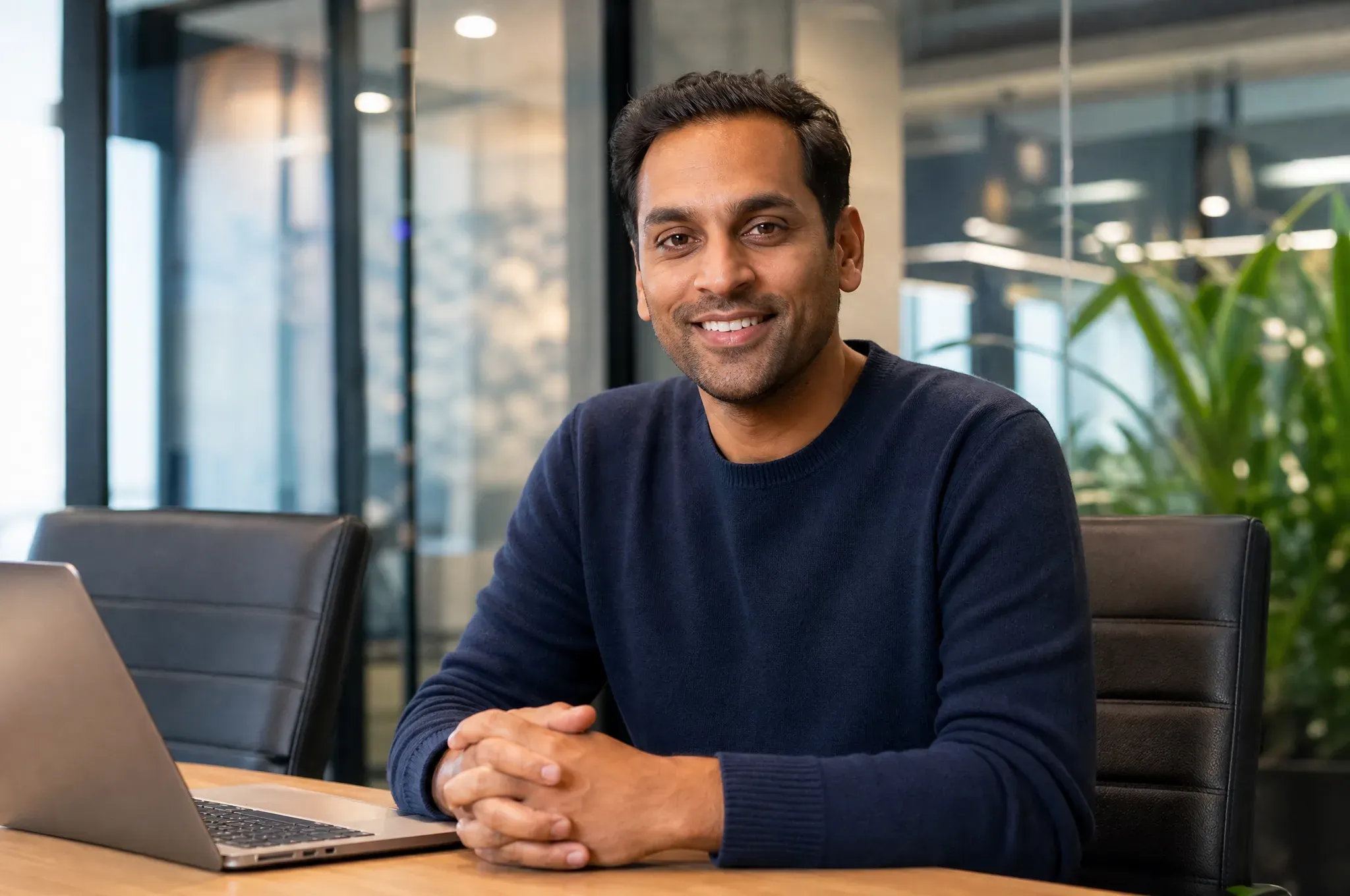 A smiling SaaS founder sits at a conference table with his hands clasped beside an open laptop, a potted plant in the background.