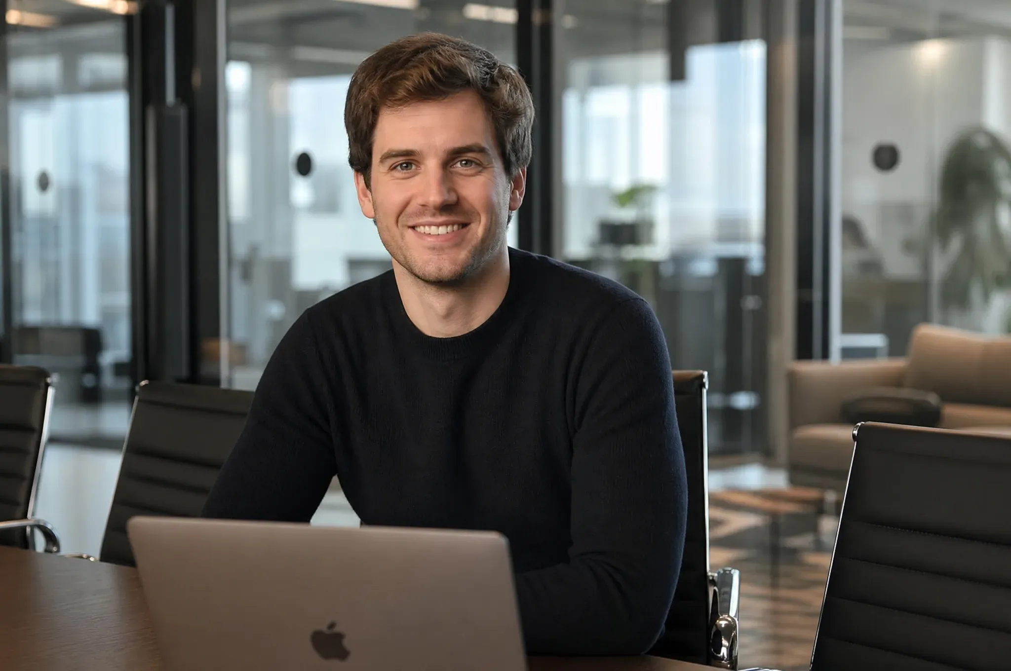 A SaaS startup founder in a black sweater smiles at the camera while seated behind an open laptop in a modern office lounge.