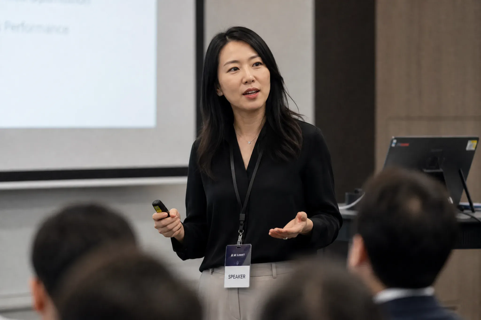 A conference speaker wearing a lanyard badge gestures while addressing an audience, with presentation slides and a laptop monitor beside her.